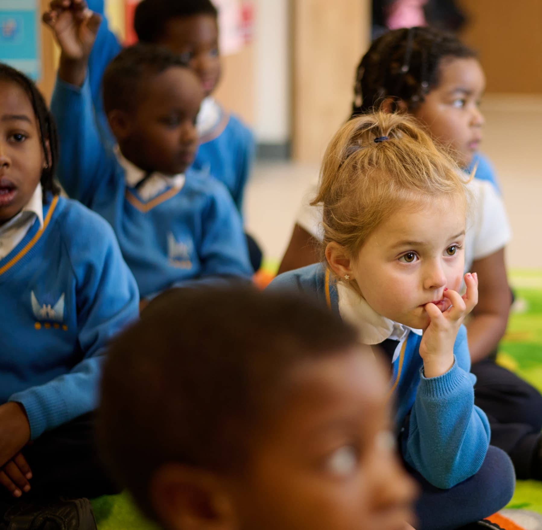 Children sitting on the floor. A child has their hand raised.